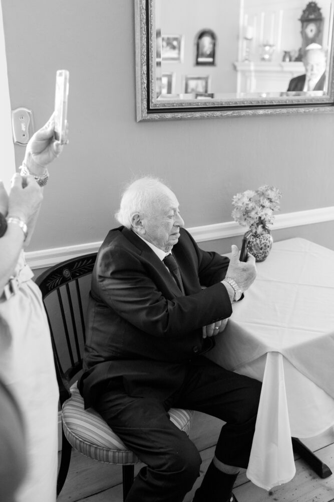 Older man in a suit sits at a dining table taking a selfie with his phone while another person nearby photographs him vase of flowers on the table