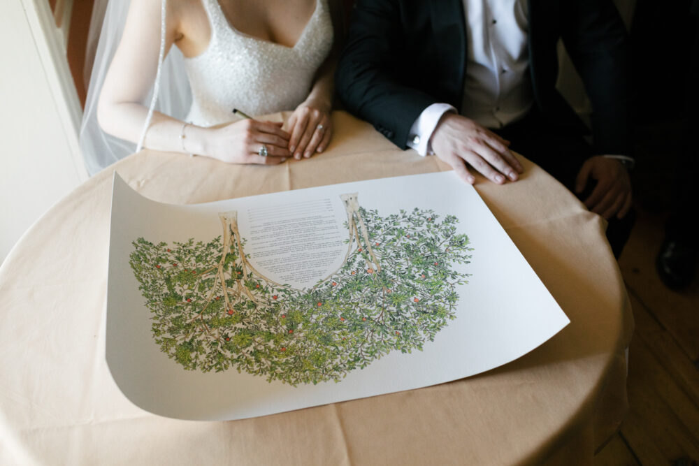 Bride and groom sign a wedding certificate with a detailed leafy tree illustration on a beige table surface
