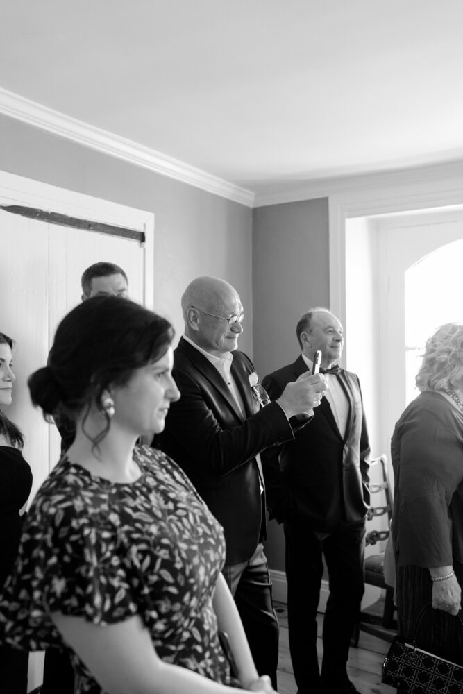 Group of formally dressed adults in a room a bald man with glasses lifts a phone to take a picture About toasts or a ceremony scene