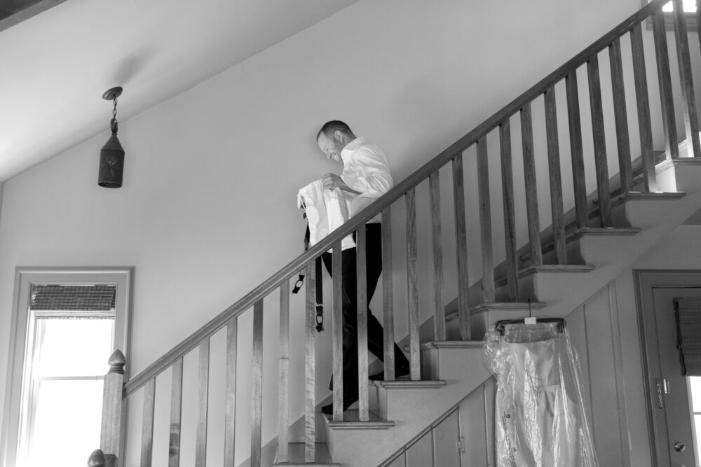 A man in a white shirt descending a wooden staircase holding a folded suit or garment inside a home
