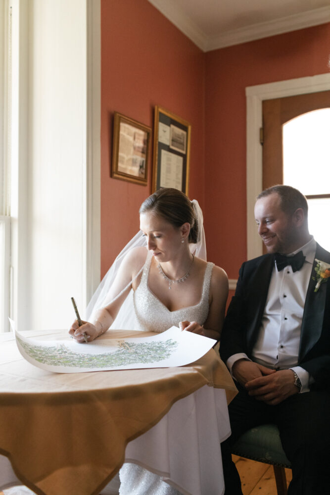 Bride in a beaded wedding dress signs a document at a table while groom in a tuxedo looks on Red walled room sunlight from window