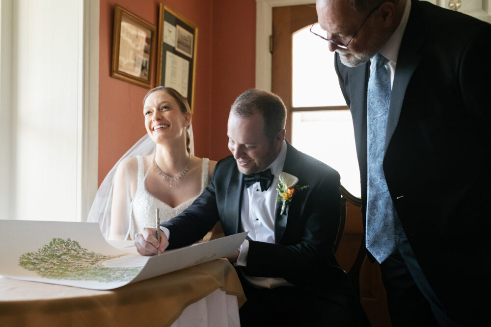Bride and groom sign a document at a table smiling as the groom signs the marriage certificate with a man standing nearby in a suit