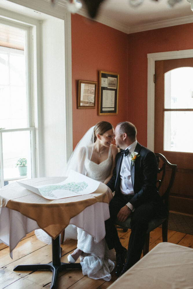 Bride in a white wedding gown and veil sits with her groom in a tux at a round table by a window sharing a smile