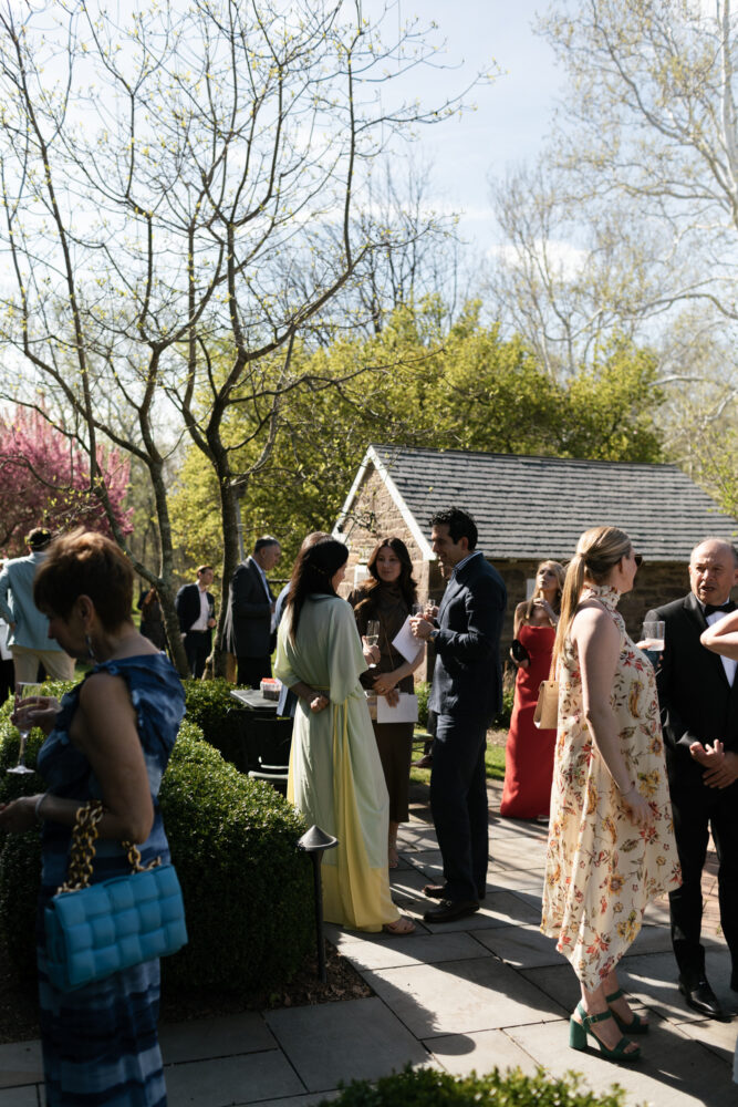 Group of well dressed guests mingling outdoors at a sunny garden party holding drinks near a brick shed and trimmed hedges