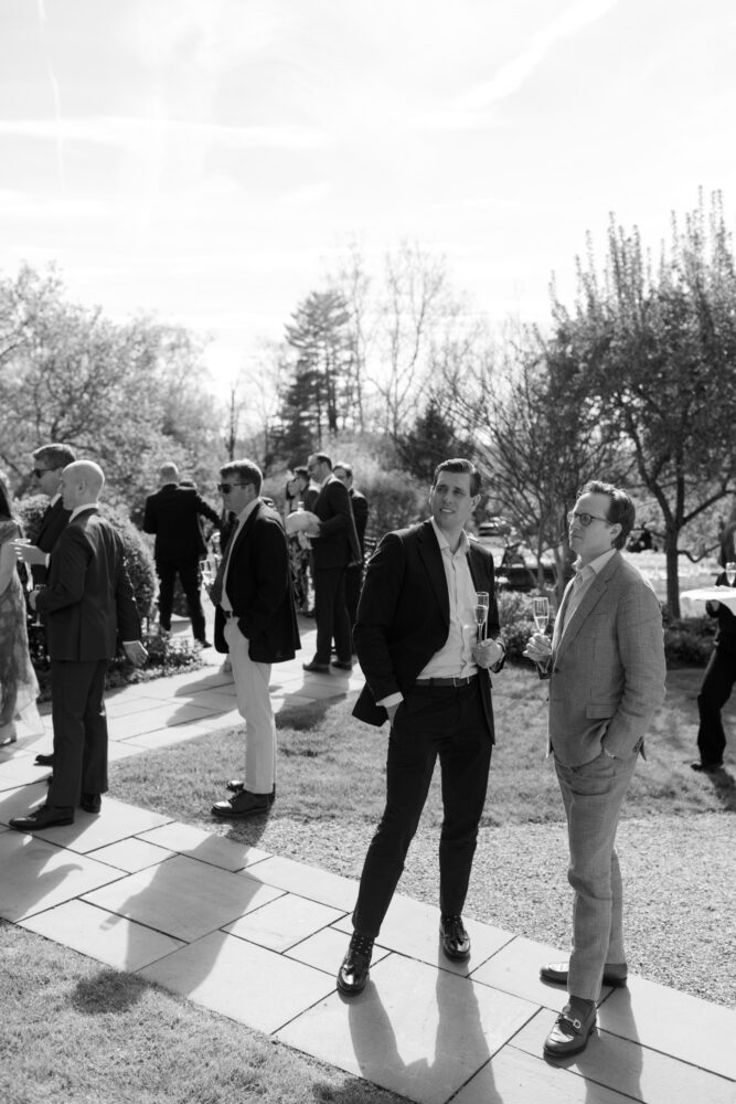 A group of well dressed men at an outdoor social event some holding champagne glasses standing on a stone patio with trees in the background