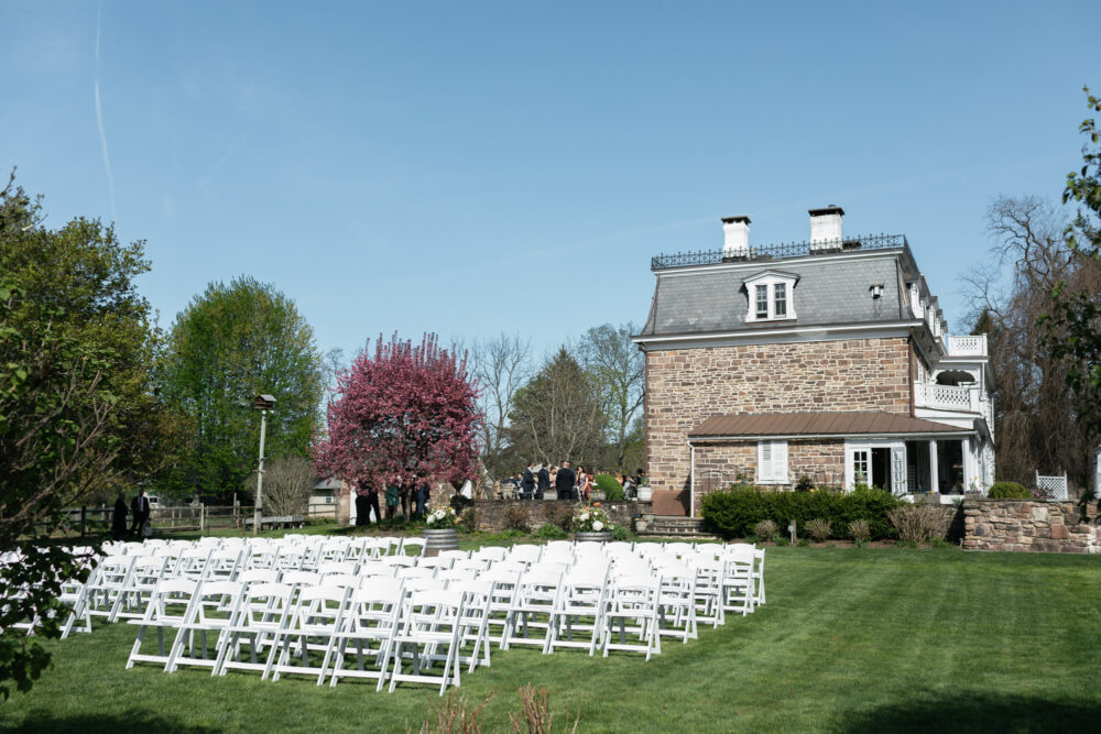 Outdoor wedding setup rows of white folding chairs on a green lawn in front of a stone mansion under a clear blue sky with a pink flowering tree nearby