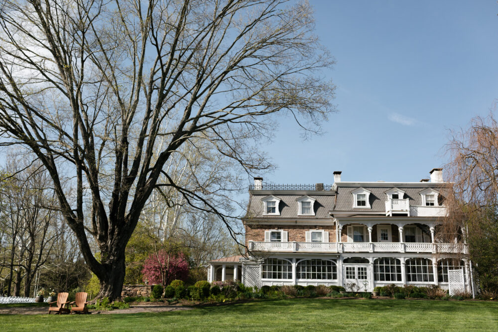Historic two story brick mansion with a wraparound porch and dormer windows set on a manicured lawn with a large leafless tree in the foreground