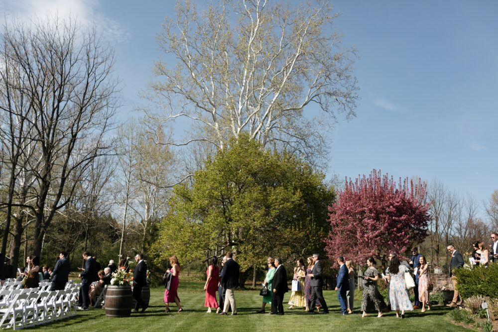 Guests walk down a grassy aisle at an outdoor wedding with white chairs on the left and blooming trees in the background