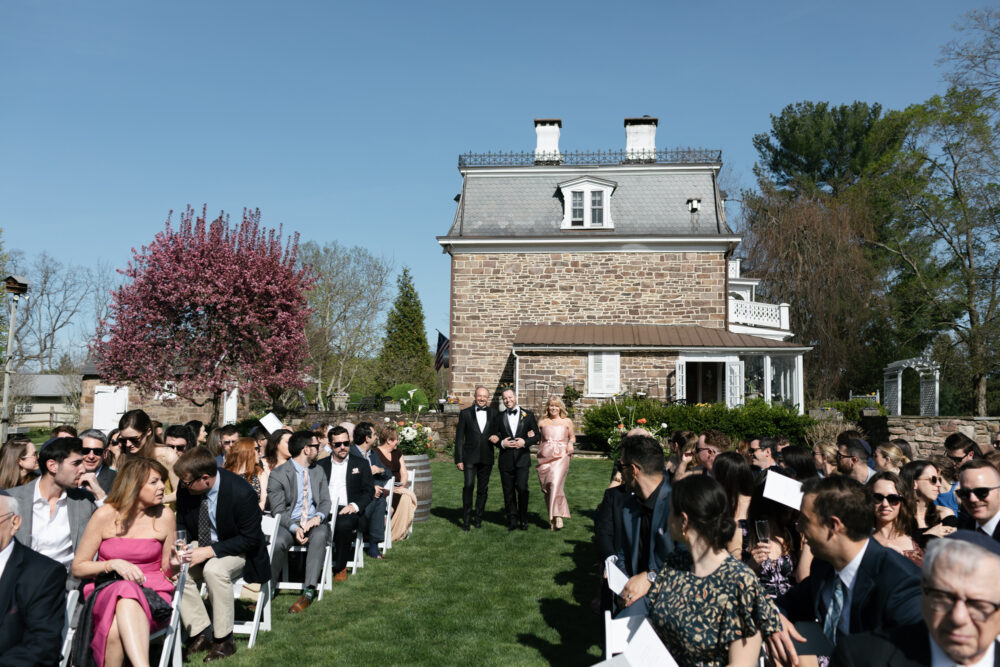 Guests seated on both sides as two men in tuxedos escort a woman in a pink gown down the aisle in front of a stone mansion