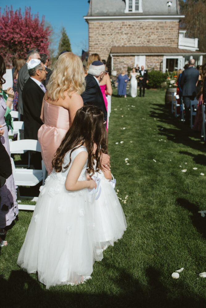 Two young girls in white dresses standing in a grassy outdoor wedding aisle as guests look toward the ceremony in the background