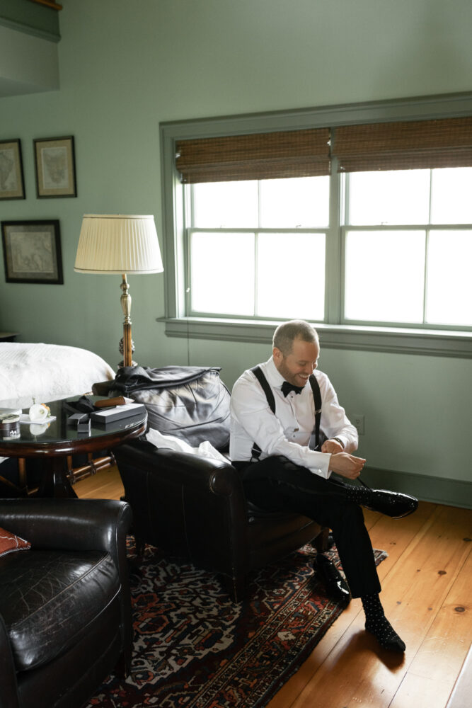 Man in a white shirt and suspenders sits on a leather chair putting on black dress shoes in a sunlit living room