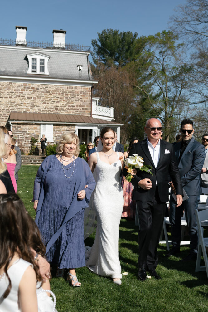 Bride in a white wedding gown walks arm in arm with a smiling woman in a blue dress escorted by a man in a black suit holding a bouquet outdoors at a sunny wedding
