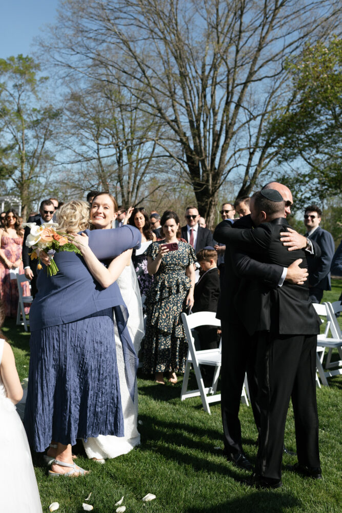Bride in a white gown hugs a woman in a blue dress at an outdoor wedding with guests smiling in the background on a sunny day in a park