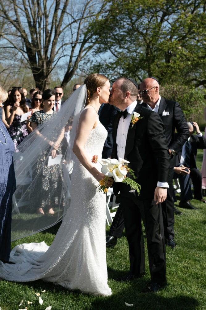 Bride in a sparkling white gown and veil kisses her groom in a black tux outdoors bouquet in hand as guests look on