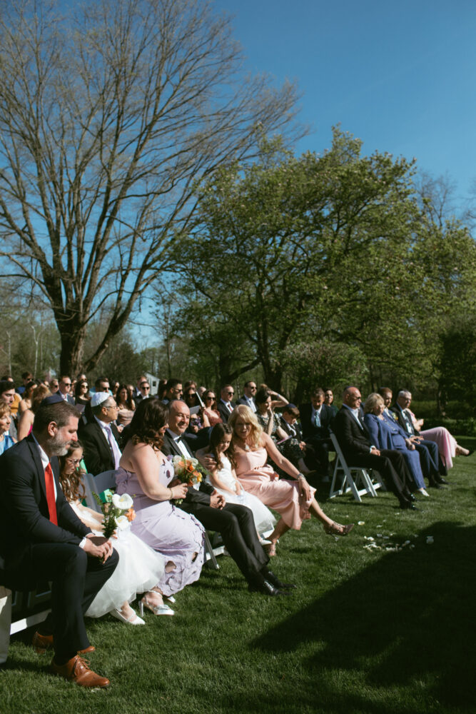 Outdoor wedding ceremony with guests seated on white chairs on a green lawn tall trees and a blue sky in the background