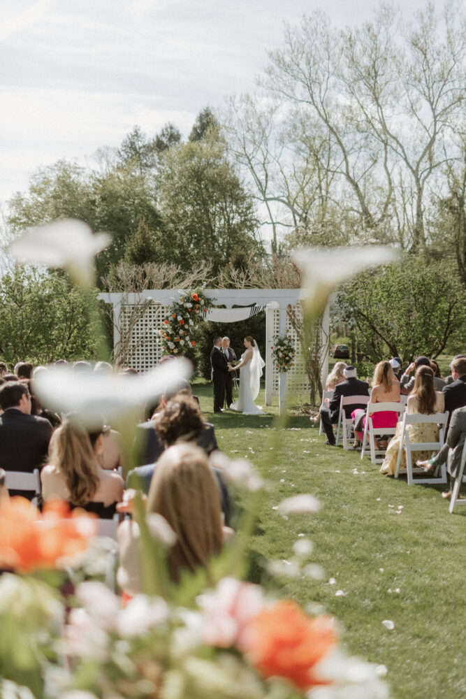 Outdoor wedding ceremony on a sunny lawn with a floral arch and guests seated in rows of white chairs