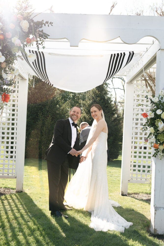 Bride and groom in formal attire smile and hold hands under a decorated outdoor wedding arch Alternative Couple in wedding dress and tuxedo stand under a striped fabric canopy outdoors on grass holding hands and smiling Alternative Smiling bride in a white gown and groom in a black suit exchange vows under a floral arch in a sunny garden Alternative Outdoor wedding scene with a bride and groom under a pergola floral decorations and bright sunlight Alternative The couples first moment after vows during an outdoor ceremony framed by a floral arch and lattice columns