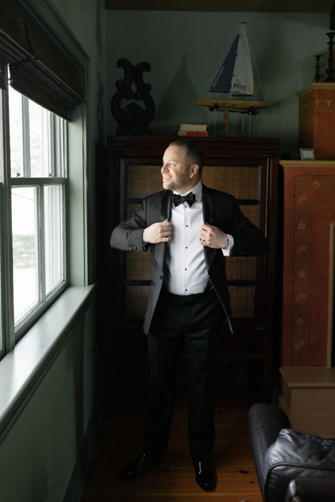 Man in a black tuxedo and bow tie stands by a window adjusting his jacket with a smile in a vintage room setting