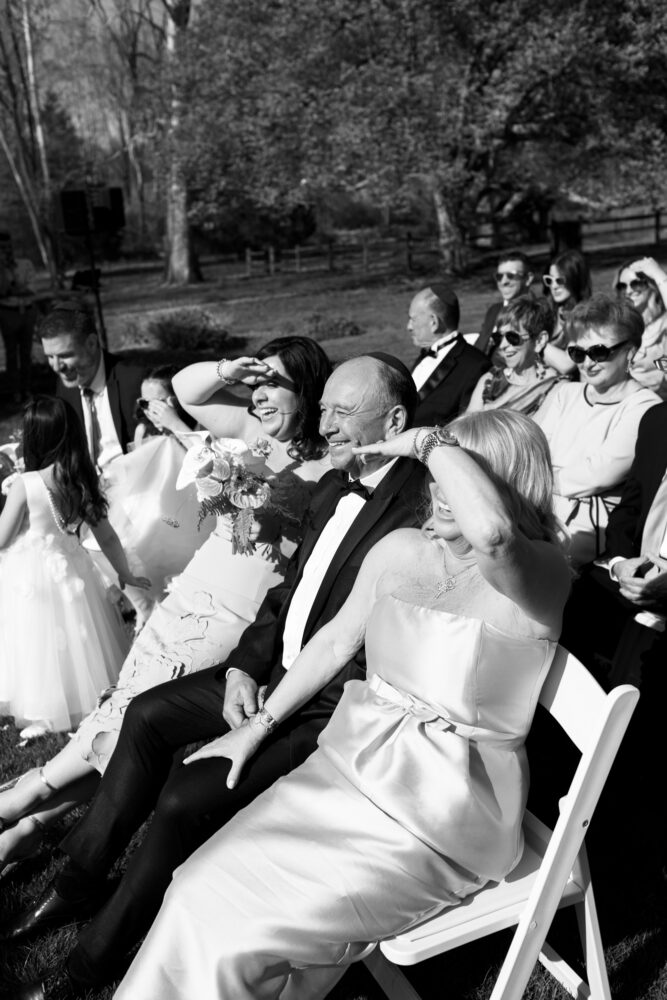 Outdoor wedding ceremony with guests in formal attire sitting on white chairs some shielding their eyes from the sun as they smile and look ahead