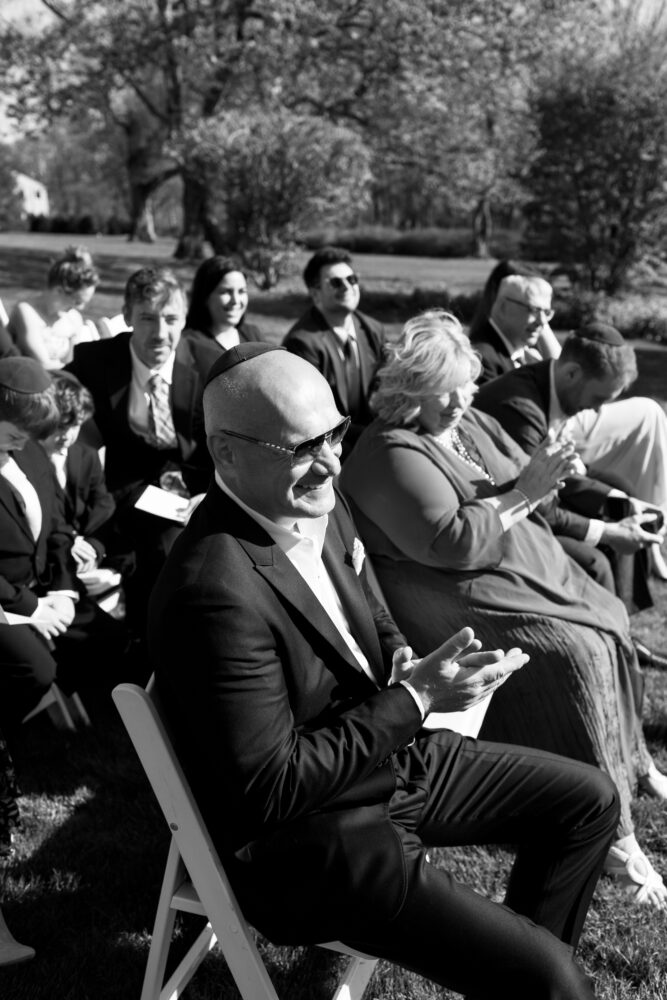 Man in a suit with a kippah sits in the foreground clapping at an outdoor wedding with guests behind him on the lawn