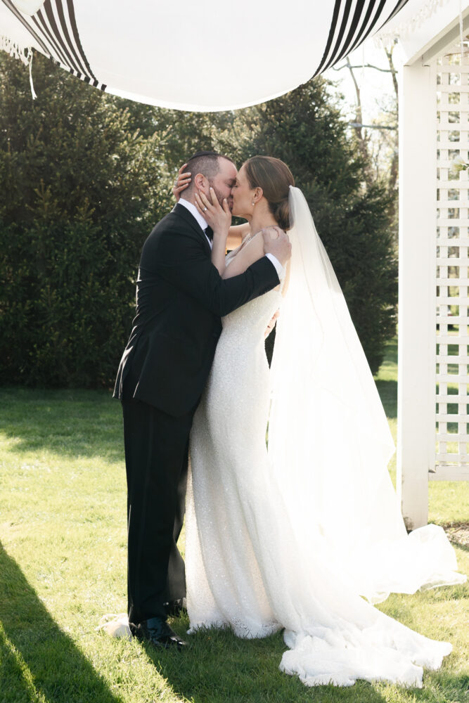 Bride and groom share a kiss outdoors under a striped canopy the bride in a sparkly white gown with a long veil