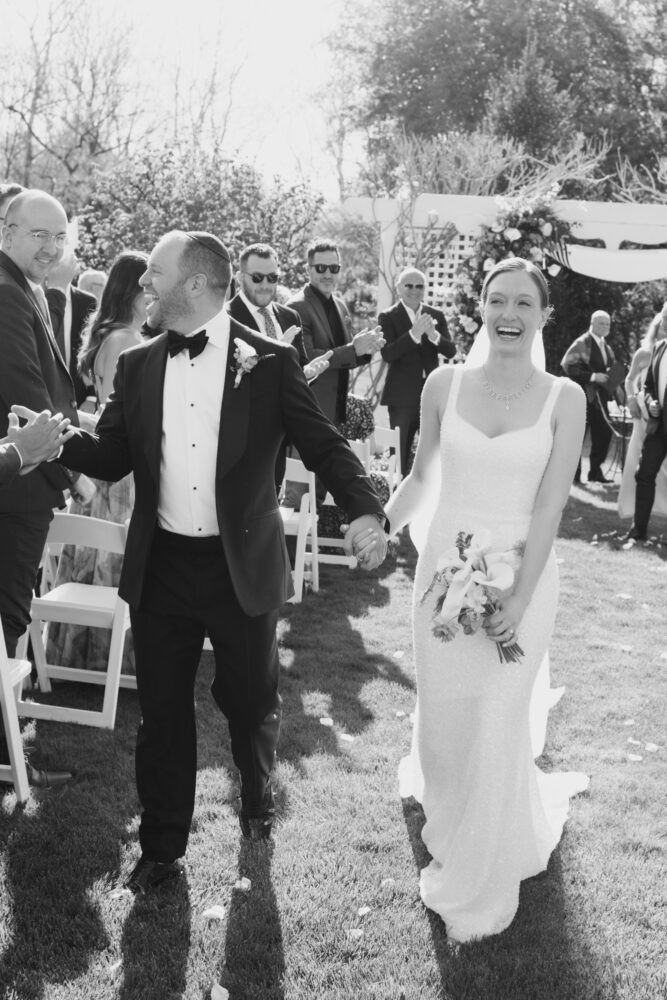 Bride and groom walk hand in hand down the aisle smiling as guests applaud outdoors at a wedding