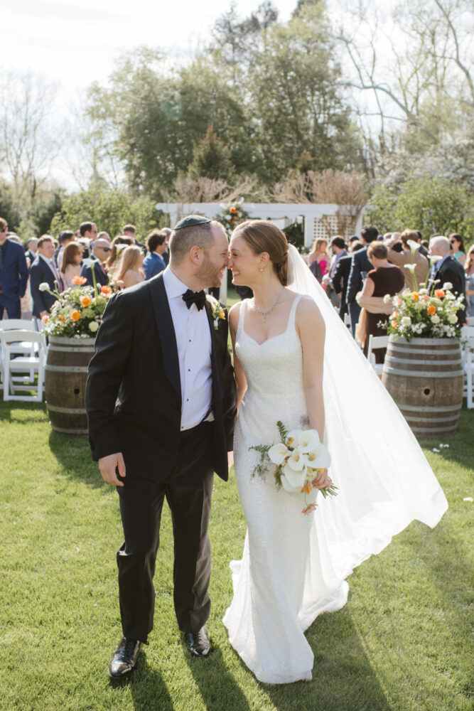 Bride in a white wedding gown with veil and bouquet of white calla lilies walks arm in arm with a groom in a black tuxedo on a sunny garden lawn as guests mingle in the background