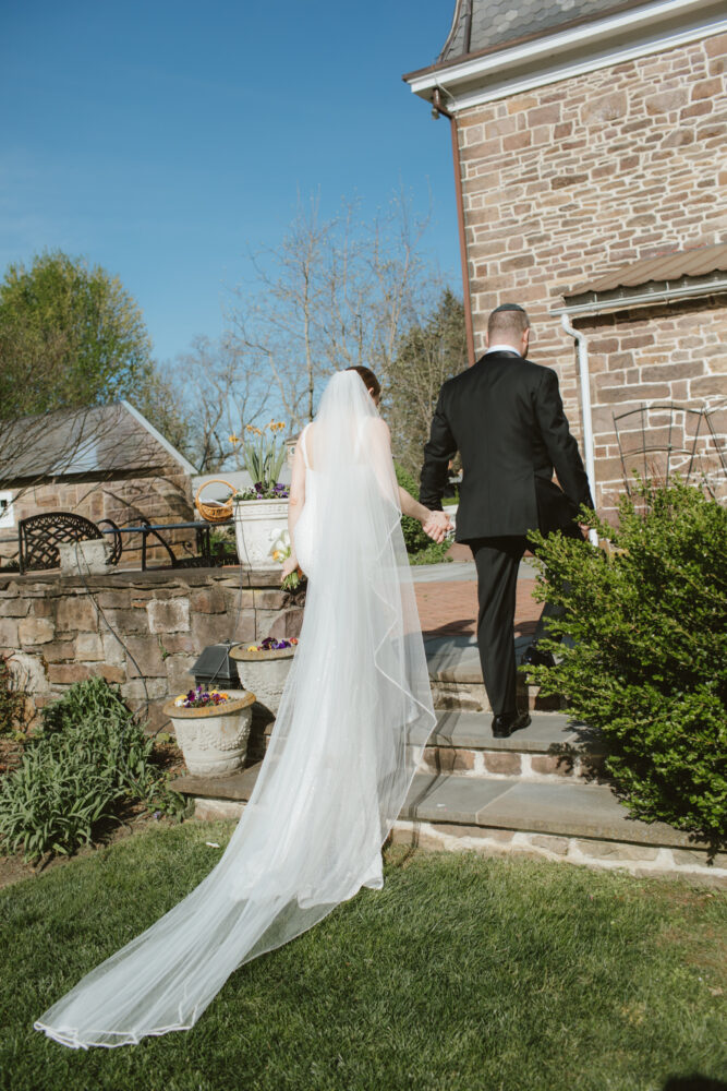 Bride in a white wedding gown and long veil walks hand in hand with a groom up stone steps beside a brick building