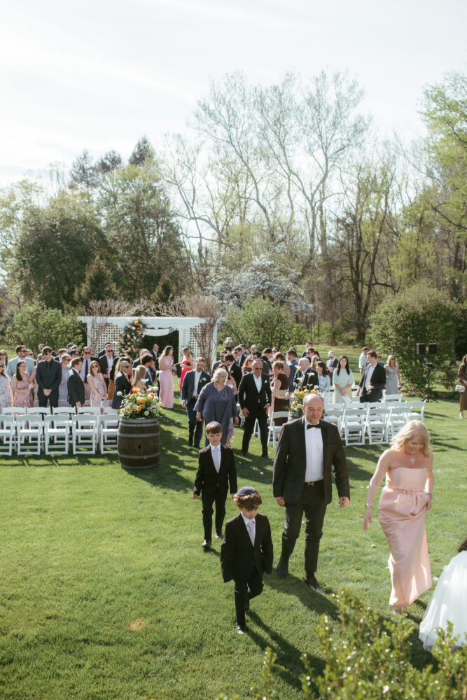Outdoor wedding ceremony on a sunny day guests in formal attire walk along a grassy aisle toward a white wedding arch with white chairs and floral arrangements on both sides