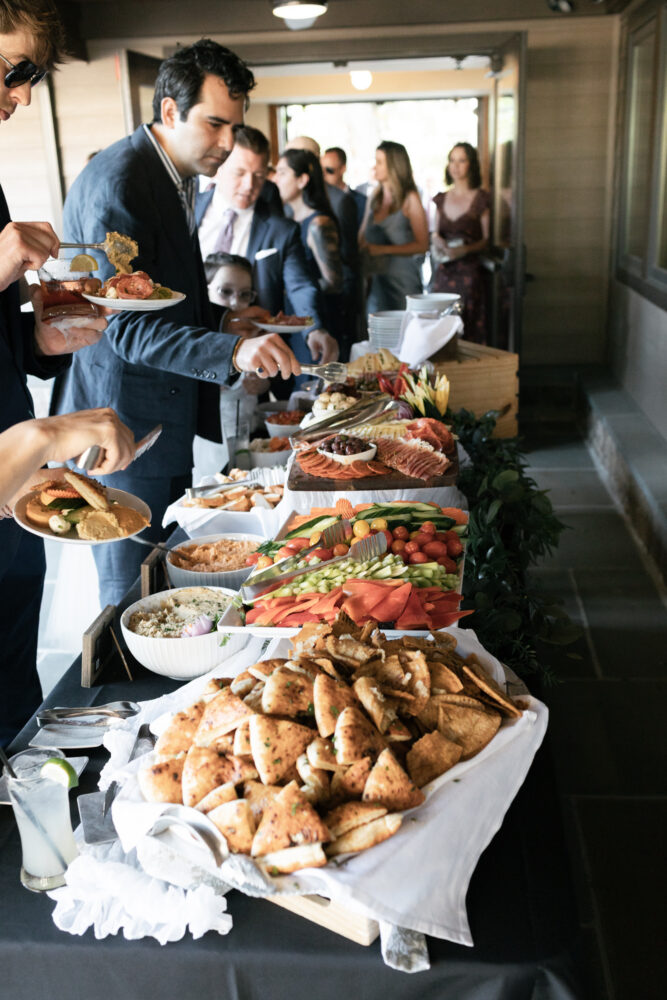 Guests serve themselves at a long buffet table filled with meats cheeses breads and colorful vegetables at a formal reception