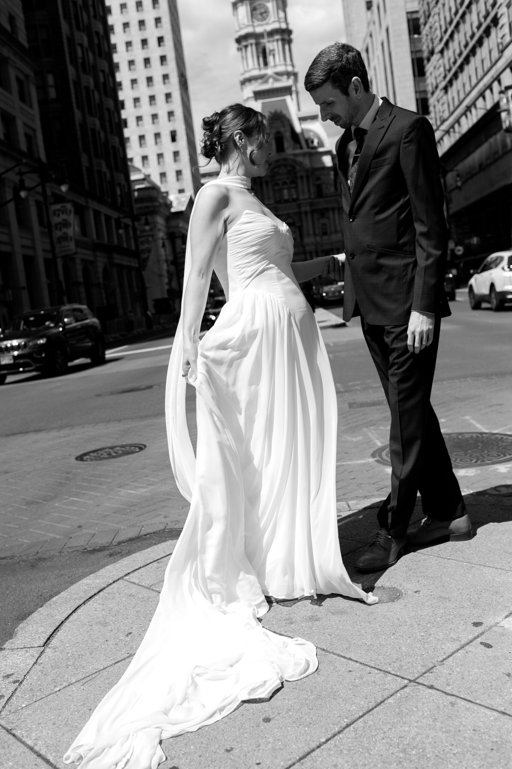 TaraBethPhotography22 Tara Beth Photography Bride in a white gown and a man in a suit hold hands on a city street sharing a tilted intimate moment between tall buildings